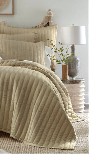 Beige quilted comforter set on a bed in a bedroom setting with pillows and a lamp.